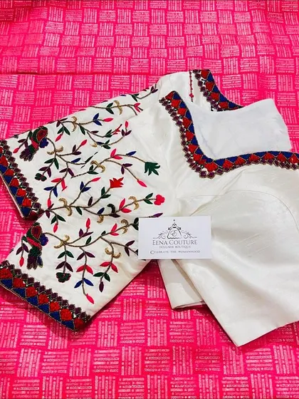 This elegant white blouse features colorful bird and floral motifs embroidered with threadwork. The neckline and sleeves are highlighted with a traditional red and blue border, perfect for pairing with a classic Kanchipuram saree.