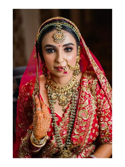 A classic bridal portrait focusing on the bride's beautiful makeup and heavy gold jewelry. Her direct gaze and traditional red lehenga exude confidence and grace.