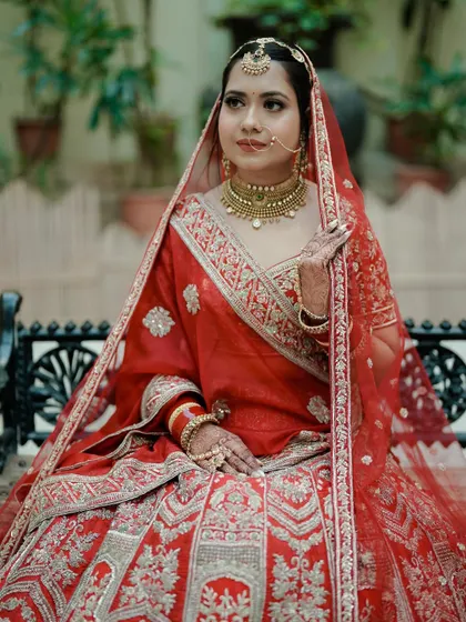 A full-length portrait of a bride in a classic red lehenga. Her seated pose on an ornate bench adds a touch of regal elegance to the shot.
