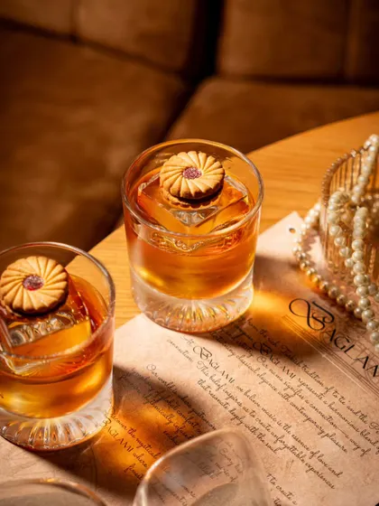 A creative shot for Baglami featuring a Jim Jam biscuit as a garnish on an Old Fashioned. This unexpected pairing, combined with moody lighting and pearls, creates a nostalgic yet sophisticated image.