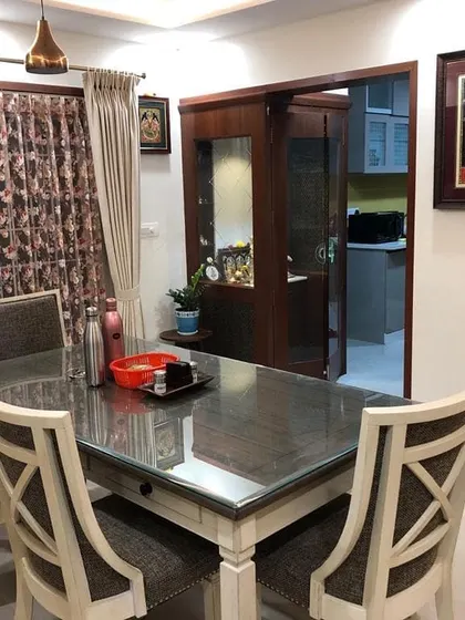 A view of a dining area that flows seamlessly towards the kitchen, showcasing an open and connected layout. The space features a modern dining table with a glass top and patterned chairs, with a glimpse of the home's pooja unit.