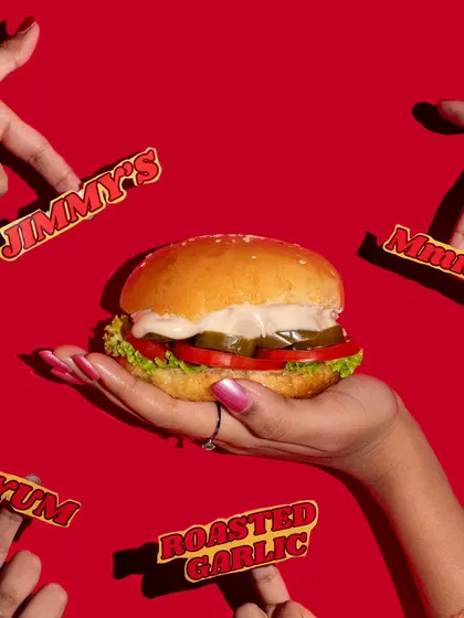 This vibrant shot of a burger against a bold red background is designed to grab attention. The composition with multiple hands pointing at the burger makes it look incredibly desirable.