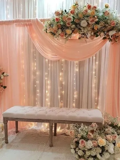 An elegant and simple photo backdrop with peach draping, fairy lights, and a floral swag.