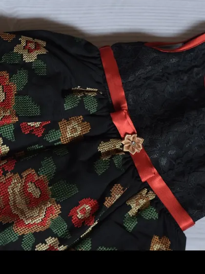 A stunning black dress featuring a beautiful cross-stitch style floral embroidery. The black lace bodice and red satin ribbon add a touch of drama and elegance.