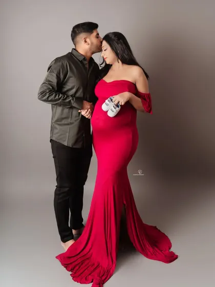 A beautiful full-length portrait of an expecting couple. The vibrant red gown and the sweet gesture of holding the baby shoes make this a classic and heartwarming maternity photo.