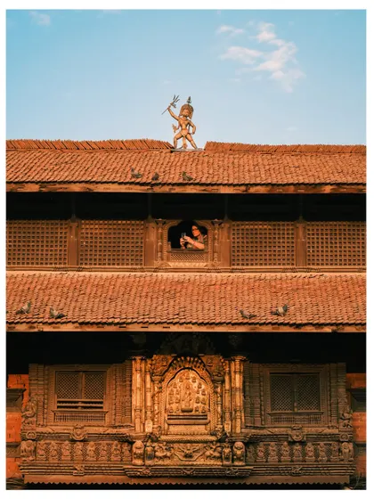 A unique view of a traditional building in Kathmandu, Nepal. The frame captures a deity statue on the roof and a person looking out from a beautifully carved wooden window.