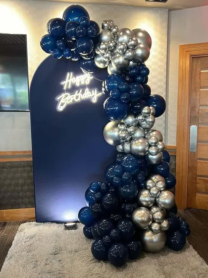 A sophisticated birthday setup with a navy blue arched backdrop and a neon sign. The balloon garland uses navy and silver chrome balloons for a sleek, modern aesthetic.