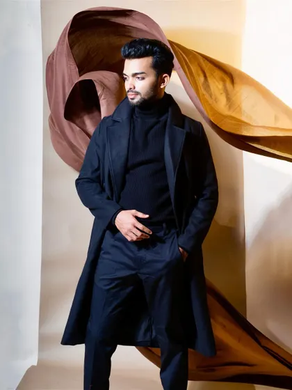 A full-length portrait of a male model in a sophisticated, dark navy trench coat and matching turtleneck. The studio shot uses draped brown and gold fabrics in the background to create an artistic, high-fashion mood for this menswear campaign.