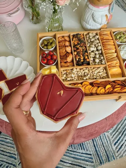 A hand holding one of our custom-designed cookies, shaped like a handbag. In the background, you can see a beautiful savoury grazing platter, showing how our sweets can complement any event.