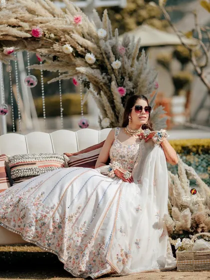 A relaxed portrait of the bride seated on a couch at her Mehendi, her elegant white lehenga and the pampas grass decor creating a chic, bohemian vibe.