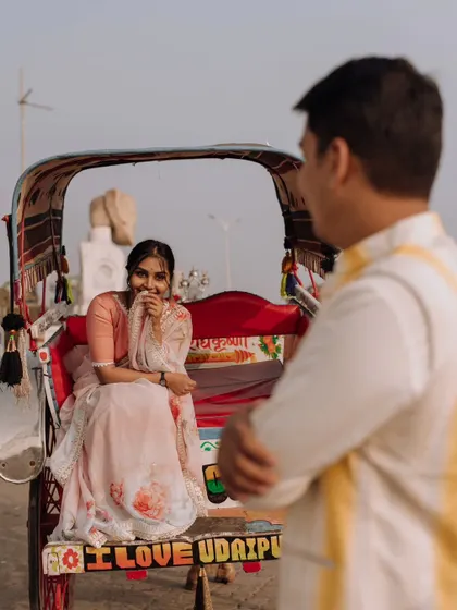 A playful, over-the-shoulder shot capturing the bride's shy smile as she sits in a colorful rickshaw, with the words "I Love Udaipur" visible.