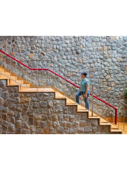 A detailed shot of the staircase against the textured stone wall. The red handrail is not just a design element; it also functions as a pipe to channel rainwater.