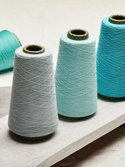 Three cones of yarn in shades of aqua and blue, displayed on a piece of rustic white wood. The composition focuses on the texture of the thread and the subtle variations in color.