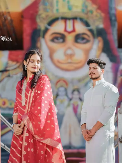 A unique and culturally rich portrait of a couple in traditional wear, posing in front of a vibrant mural of Lord Hanuman.