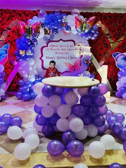 An elaborate butterfly-themed setup in a banquet hall, with a red flower wall, balloon pillars, and a balloon-based cake table.