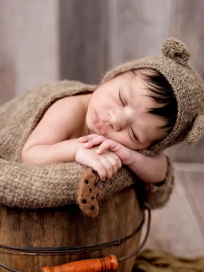 A close-up of a newborn in a bear hat, peacefully sleeping in a rustic wooden bucket. The textures of the wood and knitwear add a warm, natural feel.