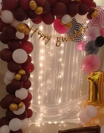 A first birthday decoration for a corner space at home. It features a balloon arch in maroon, white, and gold, with a fairy light curtain backdrop and a number "1" foil balloon.