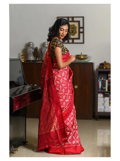 An elegant indoor portrait of a model in a red patola saree. The setting, with the piano and bookshelf, adds a touch of sophistication and context, making it a beautiful lifestyle shot.