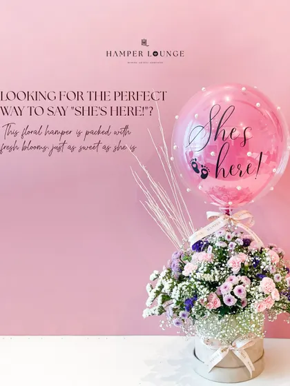 The full composition of the "She's here!" floral hamper. The soft pink background complements the delicate flowers and the pearl-adorned balloon, creating a picture-perfect birth announcement gift.