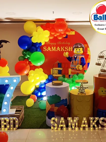 A primary-colored LEGO-themed 7th birthday party. The setup includes LEGO Batman and Emmet cutouts, LEGO-brick plinths, and a 'SAMAKSH' marquee.