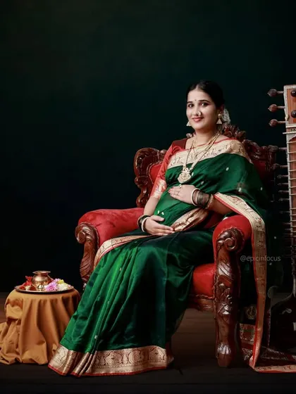 A regal portrait of a mom-to-be in a green silk saree, seated in an ornate chair with a sitar, creating a classical and artistic composition.
