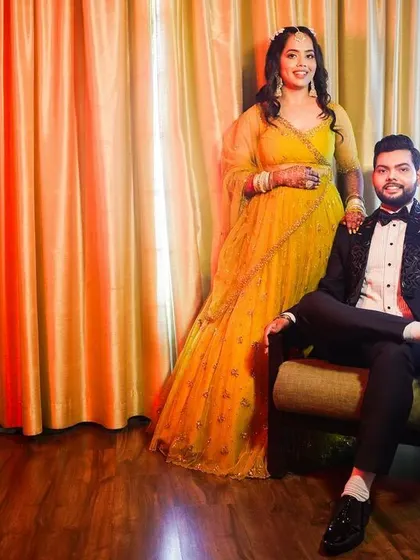 A classic couple portrait for a formal event. The groom sits while his partner stands beside him, both looking elegant and poised against a warm, glowing backdrop.