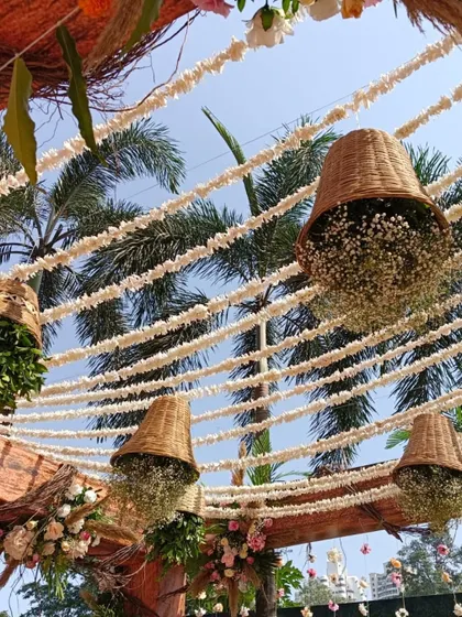 A close-up of the rustic wooden mandap, showing the detail of the hanging wicker baskets filled with baby's breath against the clear blue sky.
