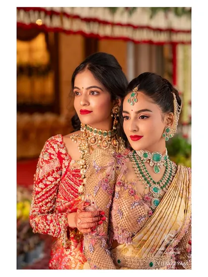 My bride Krishna with her sister, both looking absolutely regal. Krishna's makeup features a classic red lip and defined eyes, a timeless look that complements her magnificent emerald and diamond jewelry.