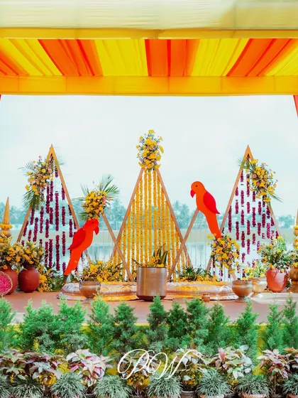 A vibrant Haldi stage with a backdrop of marigolds, geometric patterns, and parrot motifs, creating a bright and festive setting.