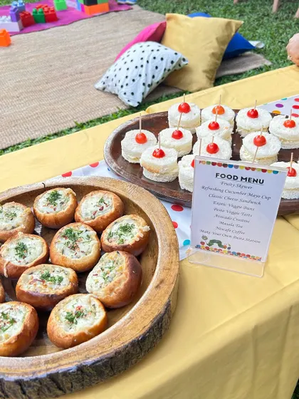A close-up of the kid-friendly food table, showing mini veggie buns and cucumber mayo sandwiches. The printed menu adds a professional and personalized touch.