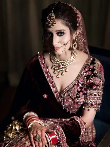 A classic, moody bridal portrait. The bride is seated in a dark room, with soft lighting that highlights her velvet lehenga and her contemplative expression, creating a very cinematic feel.