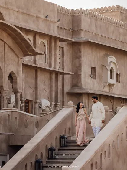 The couple descends a grand stone staircase at Fort Barwara, holding hands in a quiet, elegant moment.