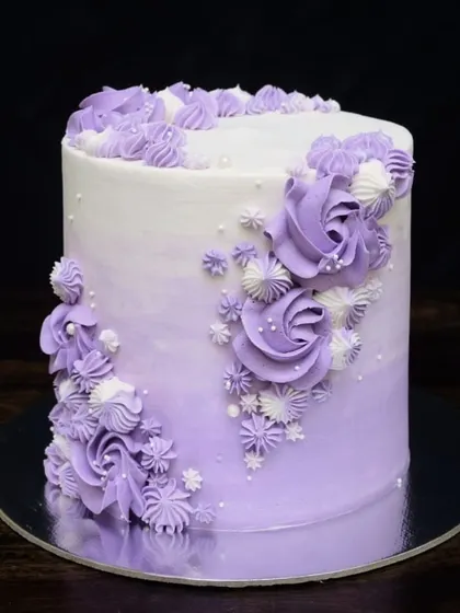 An elegant, tall egg-free cake with a purple ombre finish. The design features a cascade of beautifully piped buttercream roses and swirls in shades of purple and white.