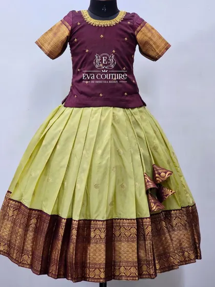 An elegant and understated pattu pavadai. The simple maroon blouse is paired with a lime green pleated skirt, both featuring a matching traditional woven border.