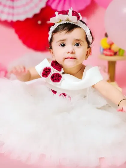 A beautiful baby girl in a white dress with red floral details, ready for her first birthday celebration.