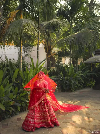 A stunning shot of the bride in her flowing red lehenga, her veil catching the light as she stands amidst tropical greenery.