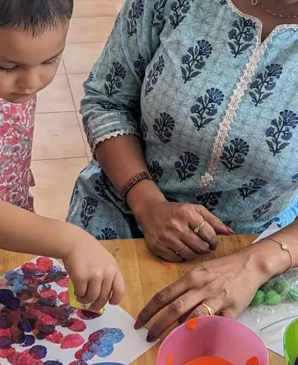 A teacher guides a child in a finger-painting activity for Holi. This sensory experience is all about exploring color and texture without the restriction of a brush.