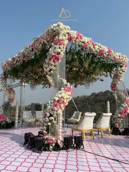 A modern and romantic open air mandap, with spiraling floral arrangements of pink and white roses on transparent pillars. The design allows for an unobstructed view of the natural surroundings.