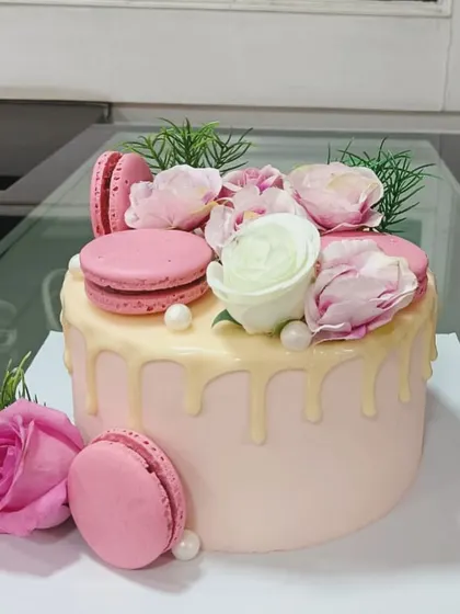 A beautiful pink drip cake, perfect for any celebration. This design features a smooth pink frosting base, a white chocolate drip, and is generously topped with fresh roses and pink macarons.
