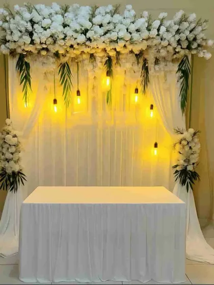 A simple and elegant backdrop for a home-based ceremony or puja, with white drapes, a floral header, and warm hanging bulbs.