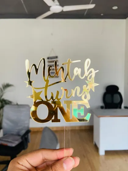 A "Mehtab turns ONE" topper held in an office setting, showing its versatility and shine in any environment.