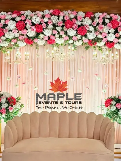 An elegant stage setup featuring a dense canopy of pink and white flowers. The soft pink drapery and classic scalloped sofa create a romantic and sophisticated setting for an engagement or reception.