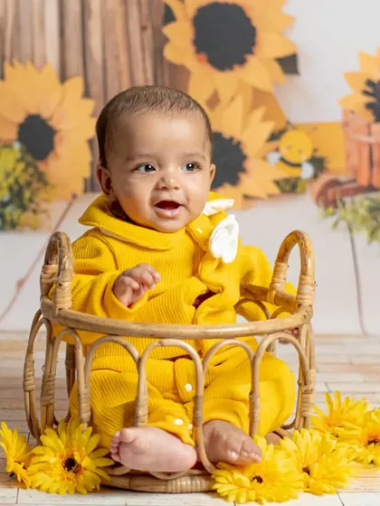 A happy baby in a yellow outfit smiles brightly from a wicker basket, part of a cheerful sunflower and bee-themed studio setup that radiates pure joy.