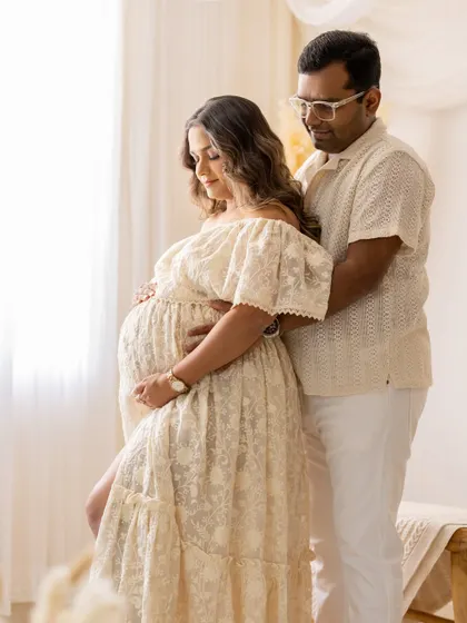 A portrait focusing on the beautiful baby bump. The couple stands together by the window, their hands gently placed on the bump, with the soft light highlighting the texture of the mother's lace dress.