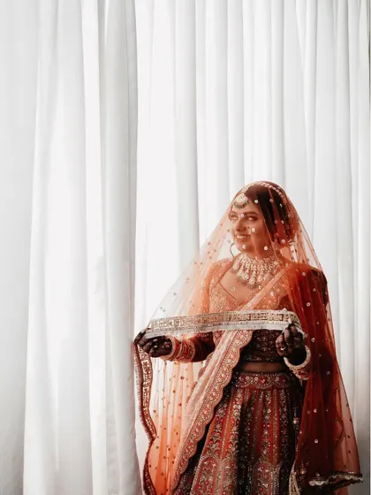 A simple, elegant portrait by a window. The bride holds her sheer red dupatta, the soft natural light beautifully illuminating her features and the details of her lehenga.