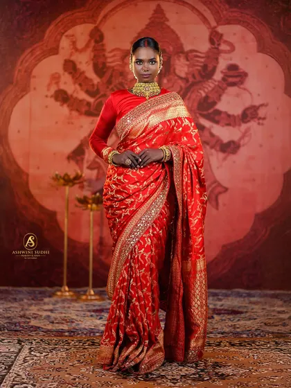 A full-length portrait that emphasizes the grandeur of the bridal ensemble. The bride stands against a traditional painted backdrop, her red saree and regal pose creating a powerful, goddess-like image.