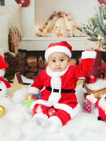 A baby dressed as Santa sits in a winter wonderland setup, complete with a snowman and Christmas tree.