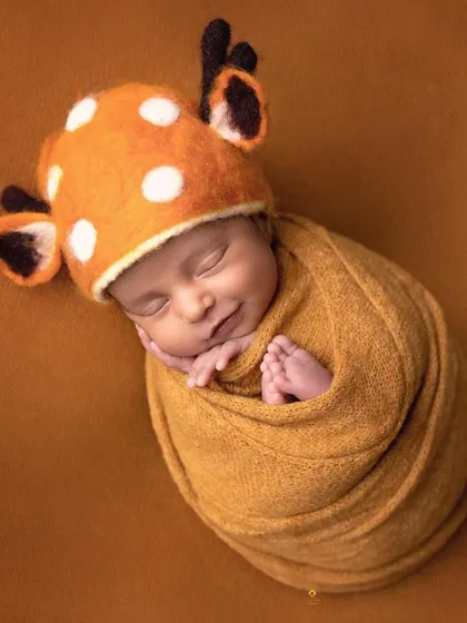 A sweet sleeping baby wearing an adorable spotted deer hat. The warm orange wrap and matching background create a cozy and whimsical woodland-themed portrait.
