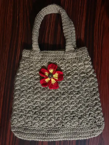 A sturdy, hand-crocheted tote bag in a neutral beige color, accented with a bright red and yellow flower for a pop of color.