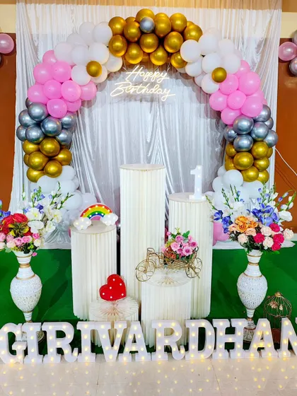 This first birthday setup uses a classic color palette of pink, gold, and silver. The decoration includes a balloon arch, flower vases, and light-up marquee letters for a grand look.